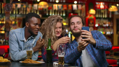 Multi-ethnic-group-of-friends-take-a-selfie-in-a-bar-and-laugh-with-a-beer-while-looking-at-photos-on-a-smartphone-screen